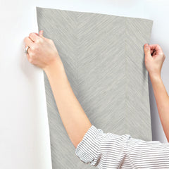 Wearing a striped long-sleeve shirt and a ring on their left hand, a person applies York Wallcoverings’ Chevron Wallpaper—a textured, light gray wallcovering with a subtle chevron pattern—to a white wall.