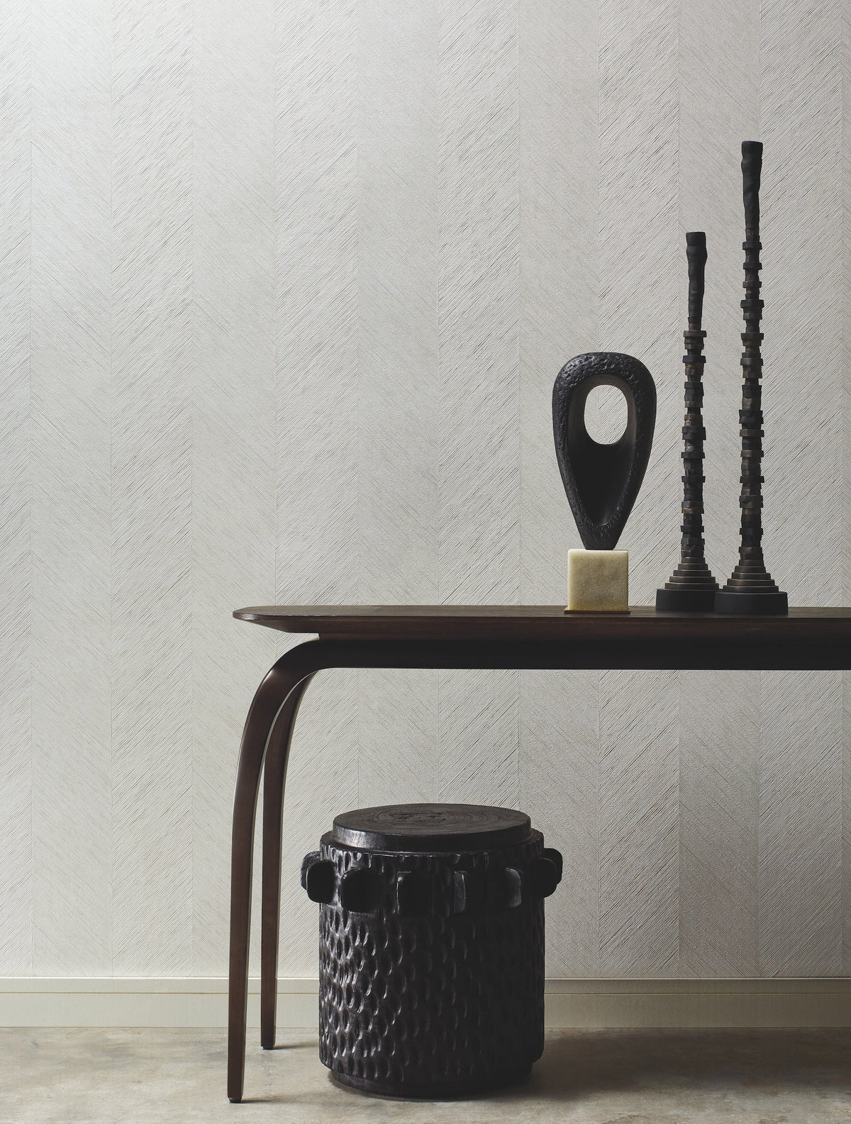 A minimalist room features a dark wooden table displaying two tall sculptural candlesticks and an abstract statue. In front sits a round, textured dark ottoman. The background is adorned with York Wallcoverings’ Chevron Wallpaper in a pale hue.