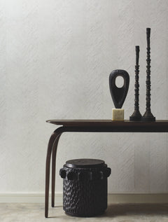 A minimalist room features a dark wooden table displaying two tall sculptural candlesticks and an abstract statue. In front sits a round, textured dark ottoman. The background is adorned with York Wallcoverings’ Chevron Wallpaper in a pale hue.