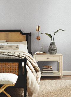 A cozy bedroom featuring a black and tan bed, beige bedding, a cream nightstand with books, a black-and-white vase with a plant, and a gold wall lamp set against York Wallcoverings’ Chevron Wallpaper.