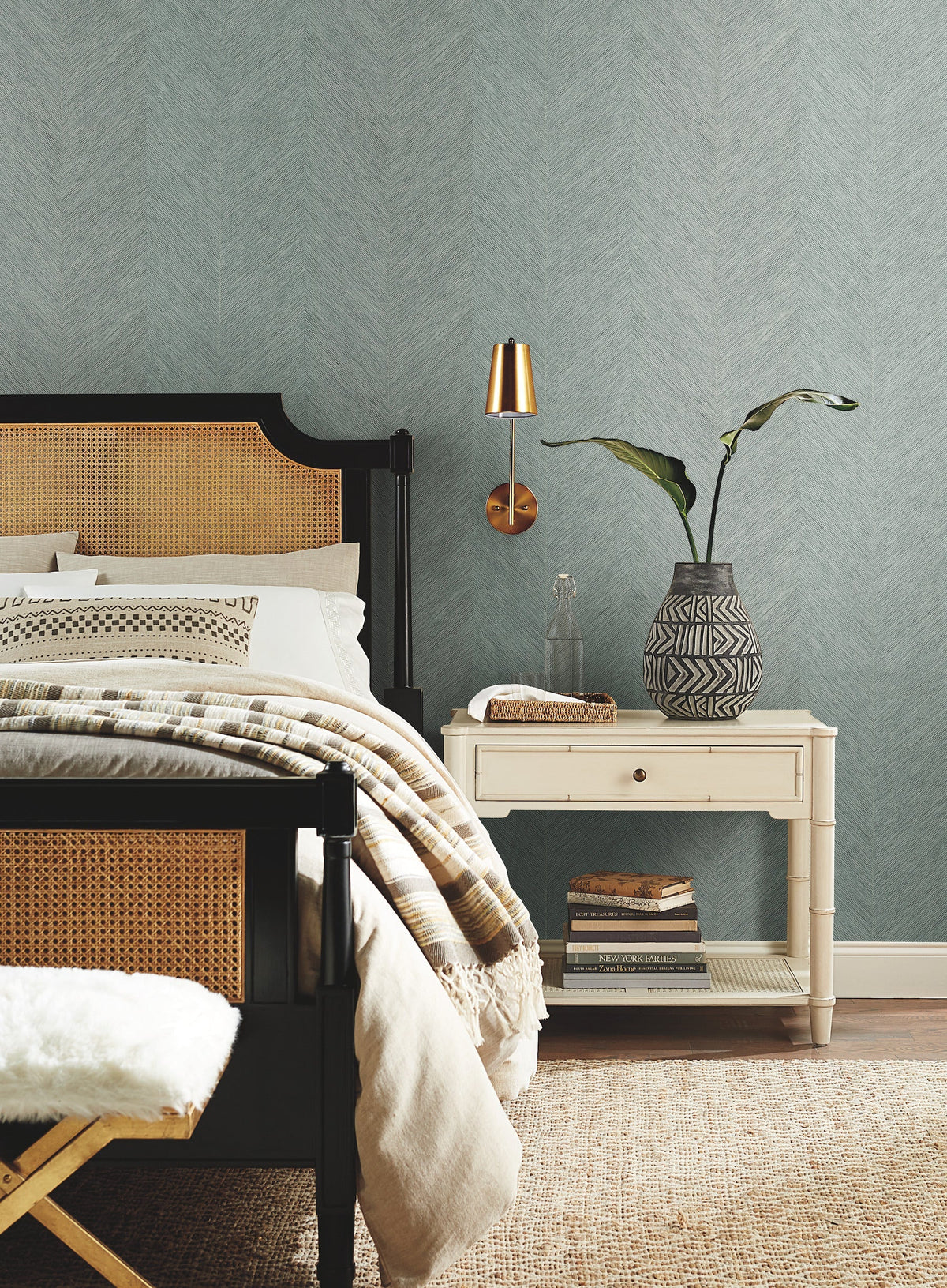 A cozy bedroom featuring York Wallcoverings’ Chevron Wallpaper in light blue, a black and tan bed with neutral bedding, a white nightstand topped with books and a decorative vase holding a plant, and accented by a gold wall lamp.