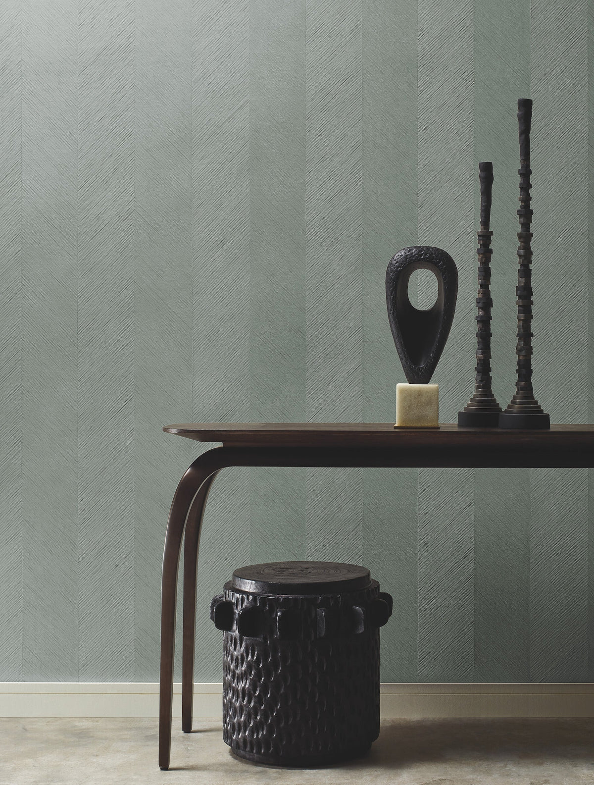 A minimalist room features a wooden table with sculptural decor, a textured black stool, and York Wallcoverings’ Chevron Wallpaper in soft green as the background.