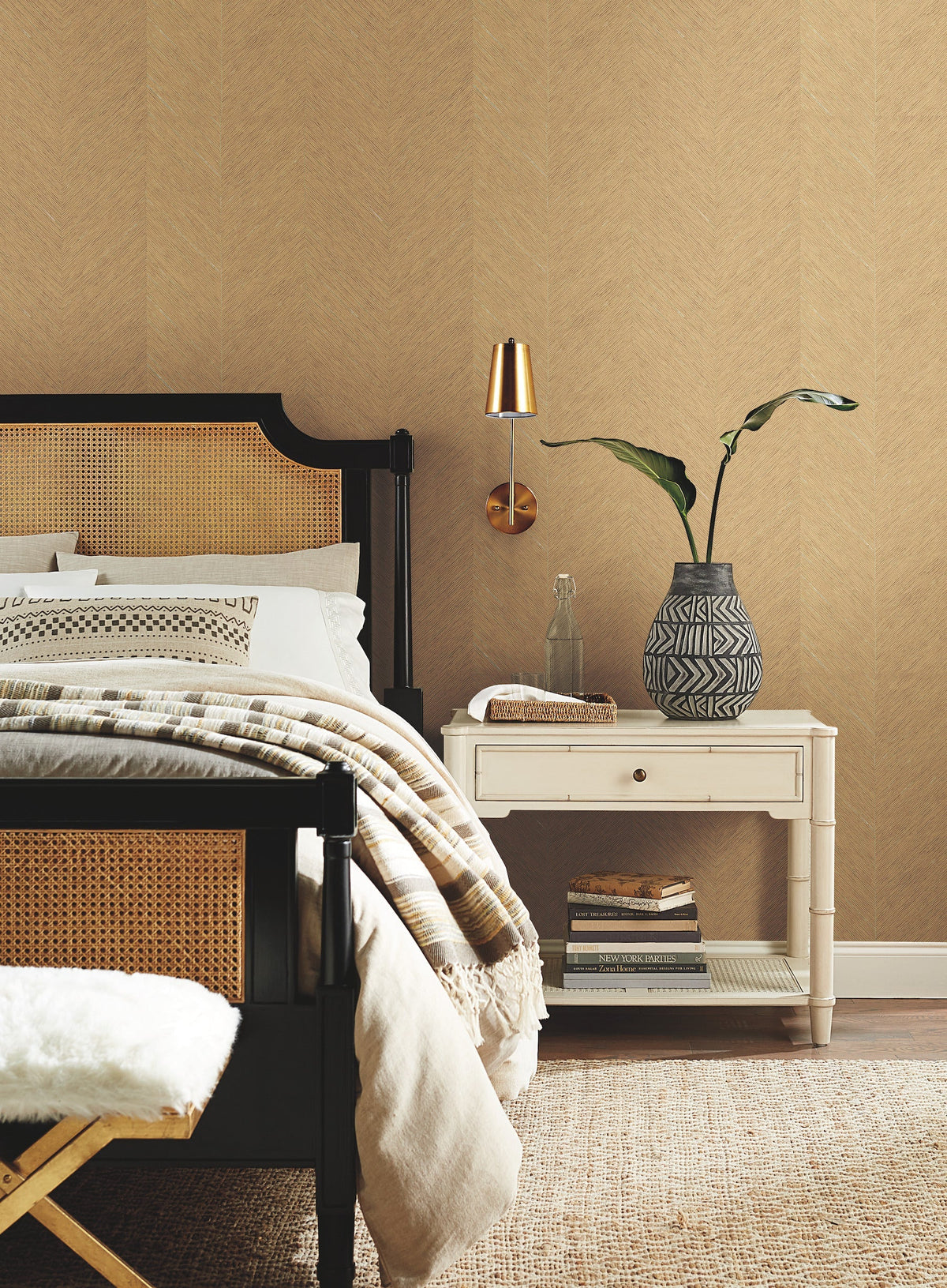 A cozy bedroom features York Wallcoverings’ Chevron Wallpaper as a backdrop, with a black and tan bed, neutral bedding, a striped throw, and a white nightstand topped with books, a glass bottle, and a patterned vase holding a leafy plant. A gold wall lamp is mounted above the nightstand.