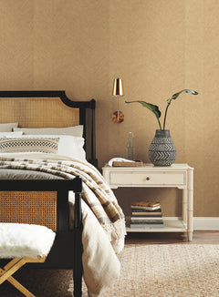 A cozy bedroom features York Wallcoverings’ Chevron Wallpaper as a backdrop, with a black and tan bed, neutral bedding, a striped throw, and a white nightstand topped with books, a glass bottle, and a patterned vase holding a leafy plant. A gold wall lamp is mounted above the nightstand.