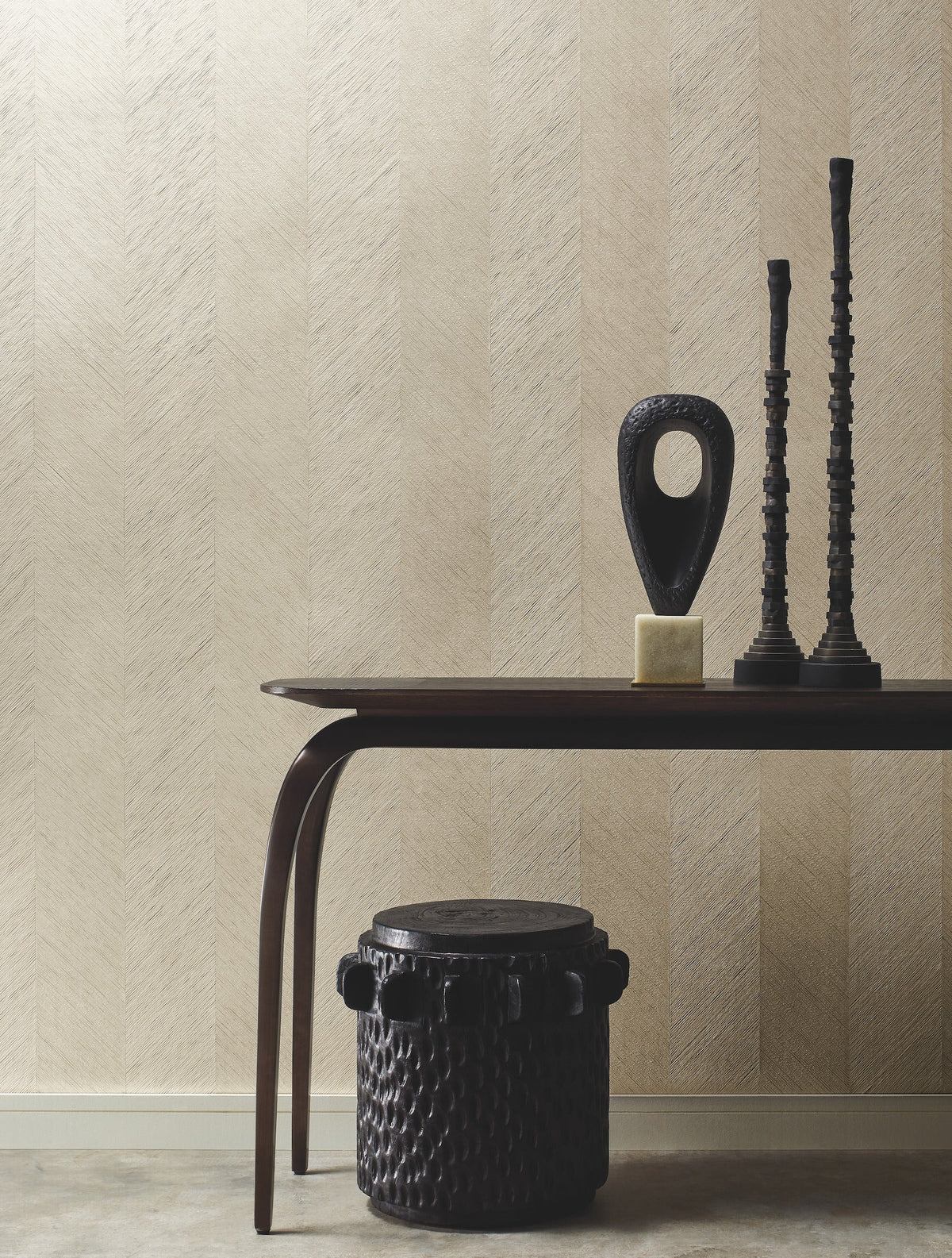 A modern table with sculptural decor and two tall candlesticks stands against a beige Chevron Wallpaper by York Wallcoverings; a dark, round, textured stool is placed underneath the table.