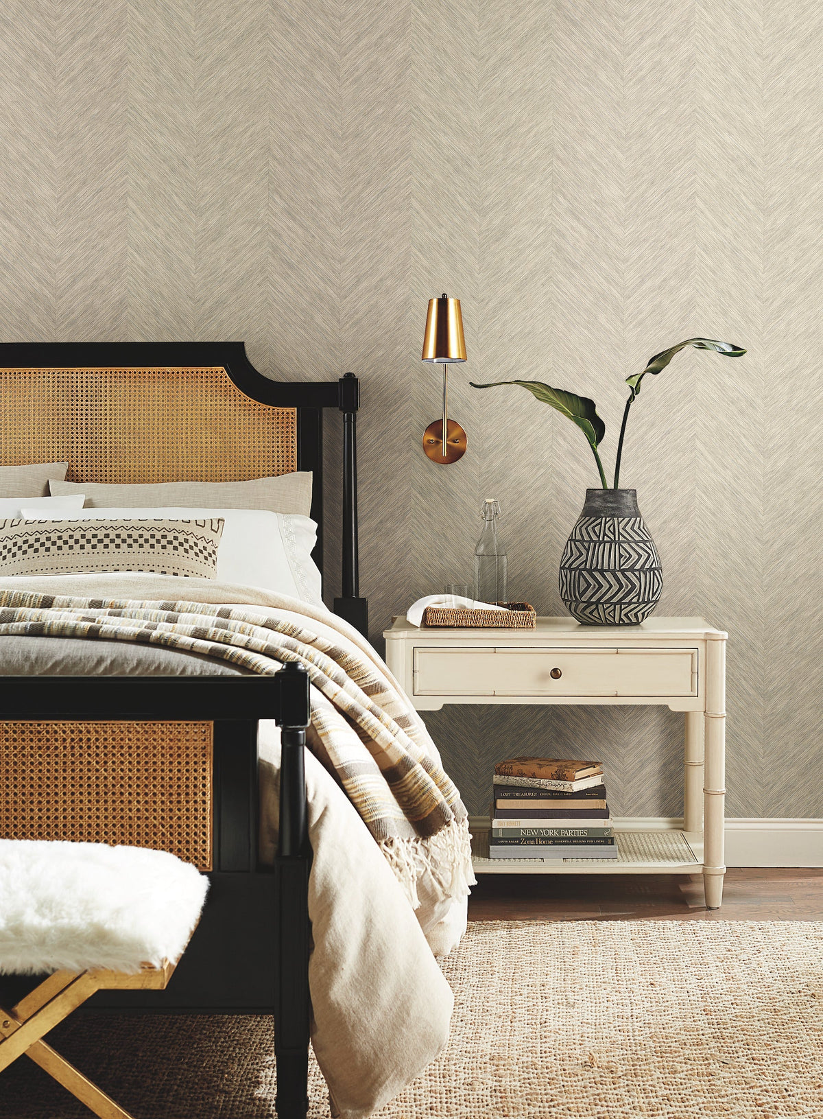 A cozy bedroom featuring a black bedframe, beige bedding, a white nightstand, a modern lamp, a patterned vase with greenery, and stacked books set against York Wallcoverings’ Chevron Wallpaper.