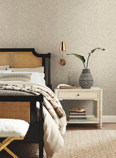 A cozy bedroom featuring a black bedframe, beige bedding, a white nightstand, a modern lamp, a patterned vase with greenery, and stacked books set against York Wallcoverings’ Chevron Wallpaper.