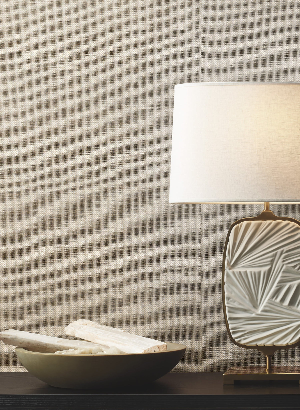 A decorative table lamp with a textured white base and a white shade sits beside a shallow bowl holding two white objects, all arranged on a dark surface in front of a beige Paper & Thread Weave Wallpaper by York Wallcoverings.