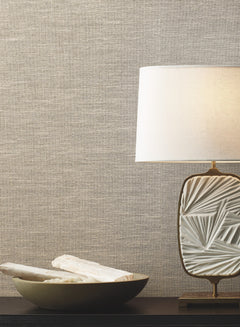 A decorative table lamp with a textured white base and a white shade sits beside a shallow bowl holding two white objects, all arranged on a dark surface in front of a beige Paper & Thread Weave Wallpaper by York Wallcoverings.
