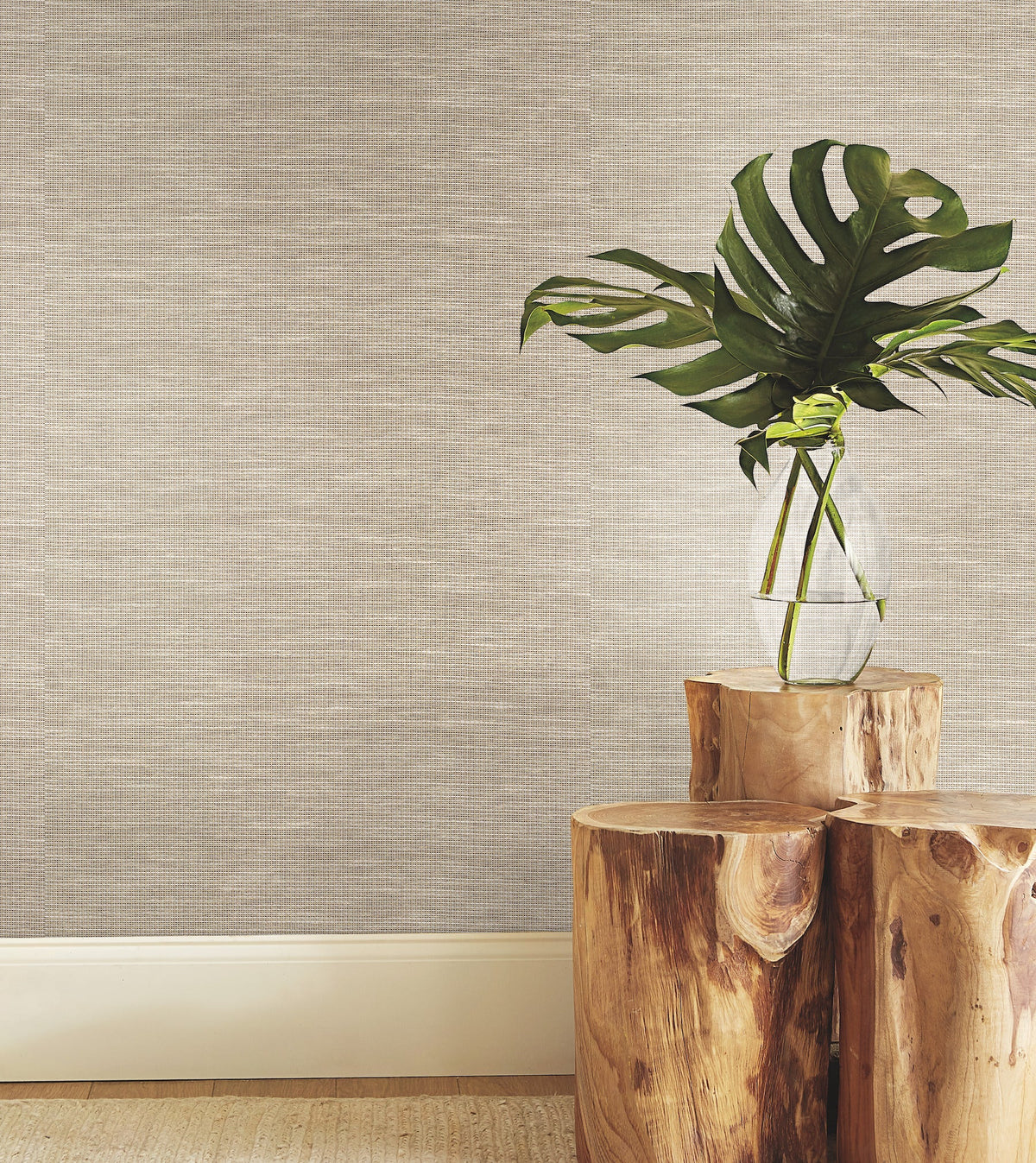 A clear vase with large green leaves sits on two stacked wooden stools against a wall finished in York Wallcoverings’ Paper & Thread Weave Wallpaper, with a light baseboard and part of a woven rug visible on the floor.