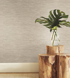 A clear vase with large green leaves sits on two stacked wooden stools against a wall finished in York Wallcoverings’ Paper & Thread Weave Wallpaper, with a light baseboard and part of a woven rug visible on the floor.