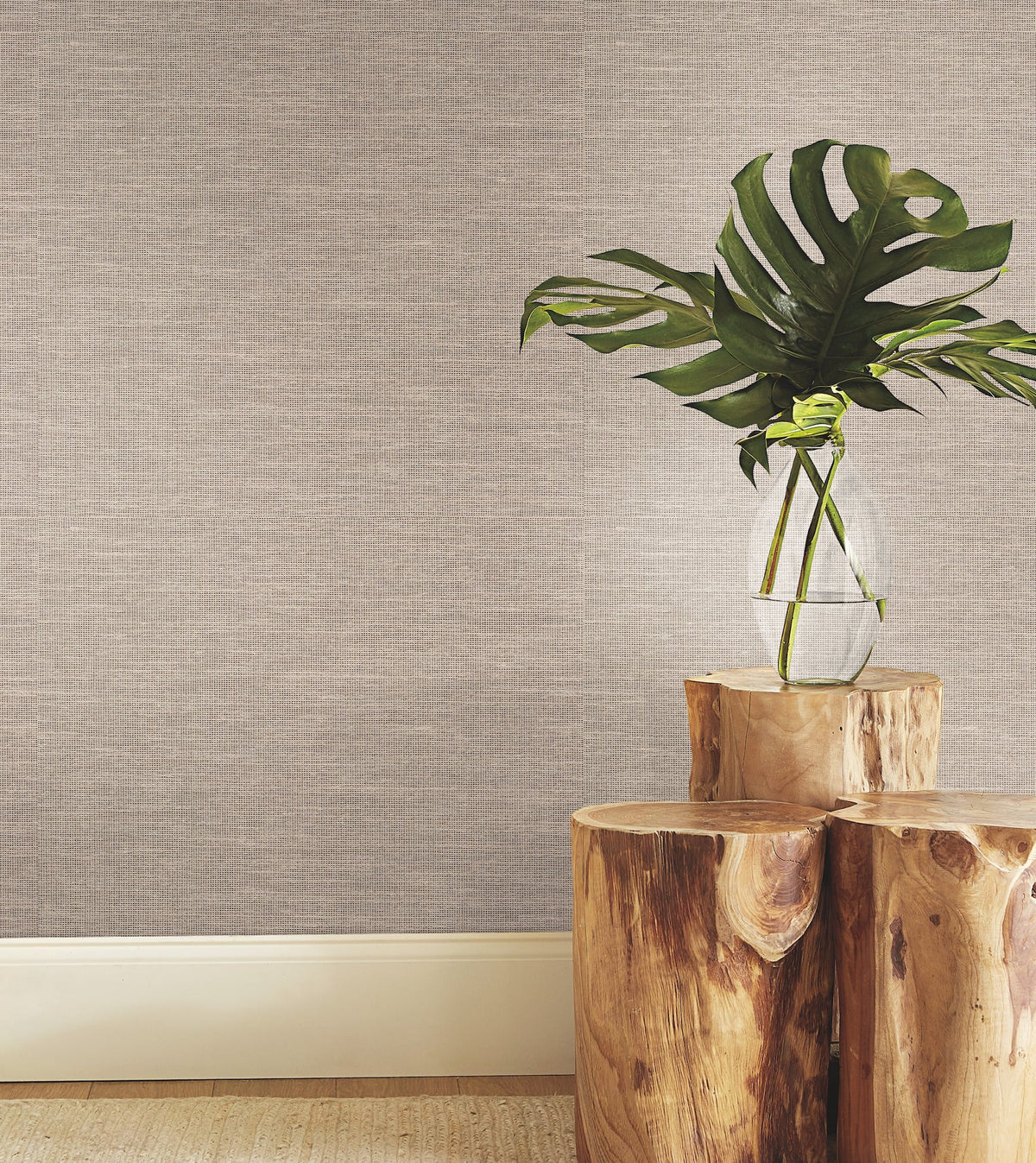 A clear glass vase with large green leaves sits on stacked wooden stumps against York Wallcoverings’ Paper & Thread Weave Wallpaper, with a white baseboard below.