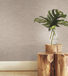 A clear glass vase with large green leaves sits on stacked wooden stumps against York Wallcoverings’ Paper & Thread Weave Wallpaper, with a white baseboard below.