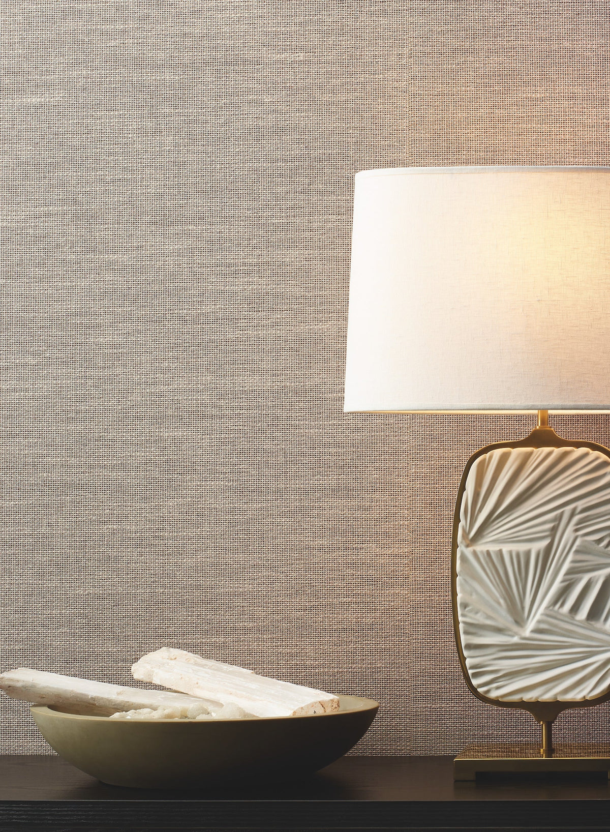 A decorative table lamp with a textured white base and a white shade sits on a black surface next to a shallow bowl holding white crystal-like stones, set against York Wallcoverings’ Paper & Thread Weave Wallpaper.