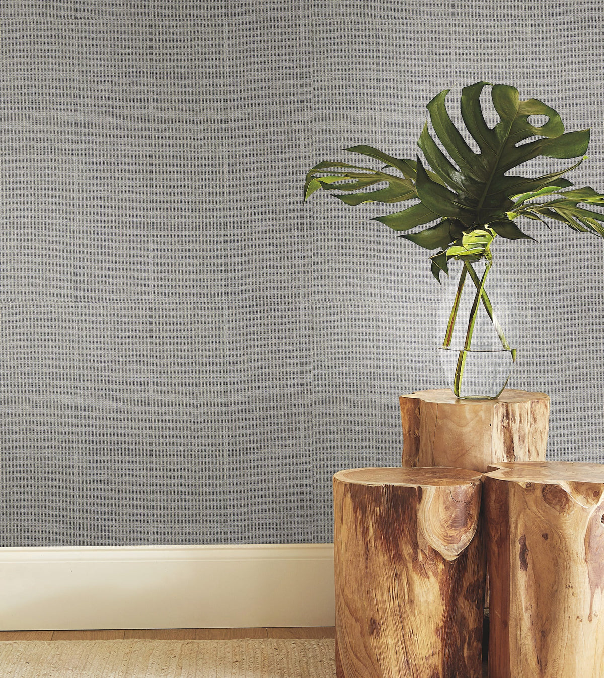 A clear glass vase with a large green monstera leaf sits on stacked natural wooden stools against York Wallcoverings’ Paper & Thread Weave Wallpaper, with a beige carpeted floor beneath.