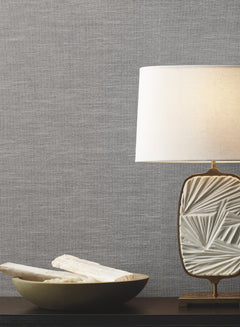 A decorative table lamp with a textured white base and a white shade sits beside a bowl with white stones on a dark surface, set against a wall adorned with York Wallcoverings’ Paper & Thread Weave Wallpaper.