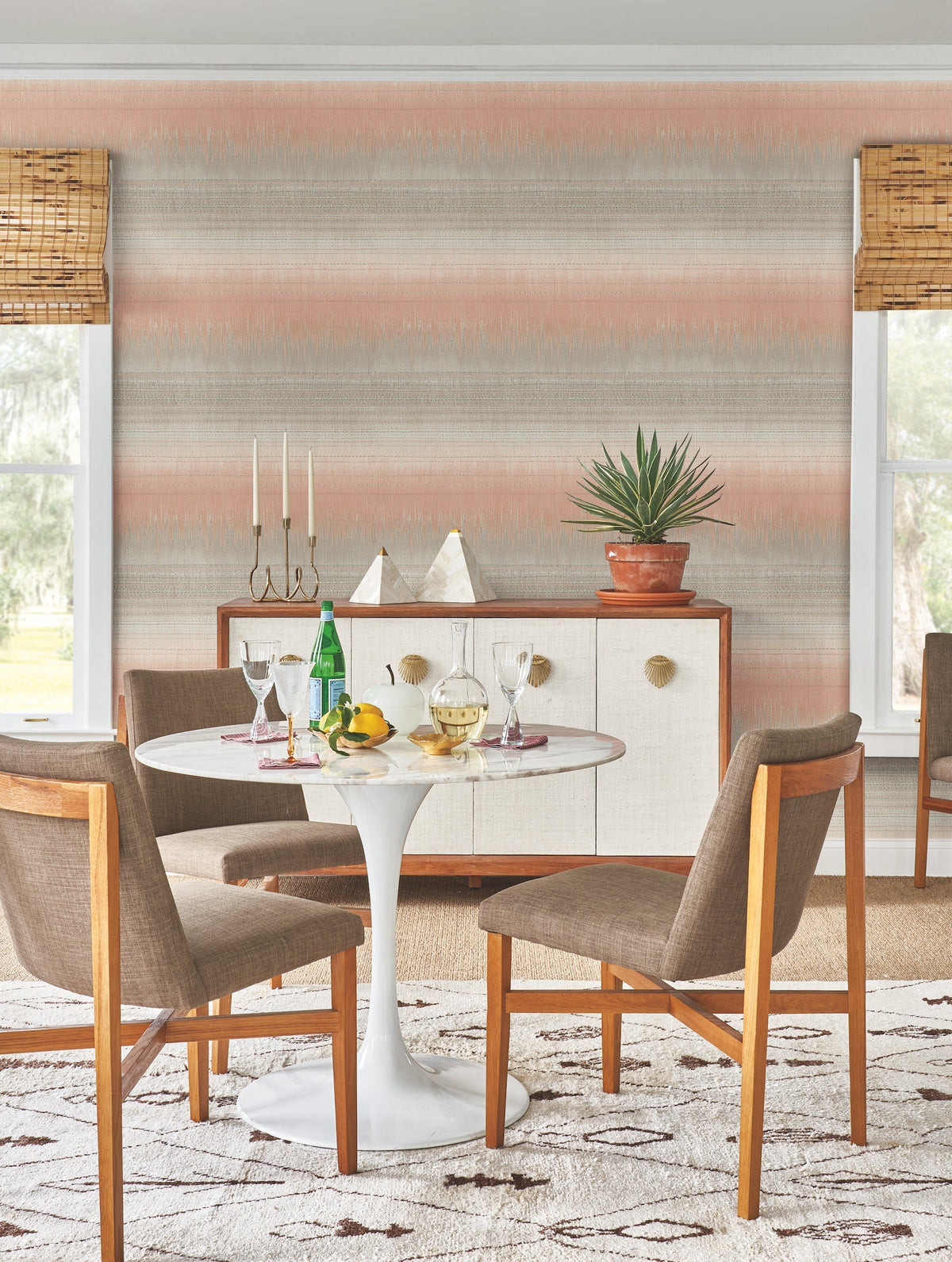 A modern dining area featuring a round white table, four brown chairs, a sideboard adorned with decor, and a potted plant. Two windows with bamboo shades bathe the room in natural light, while Desert Textile Wallpaper by York Wallcoverings adds subtle texture to the walls.