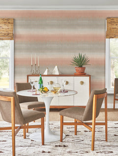 A modern dining area featuring a round white table, four brown chairs, a sideboard adorned with decor, and a potted plant. Two windows with bamboo shades bathe the room in natural light, while Desert Textile Wallpaper by York Wallcoverings adds subtle texture to the walls.