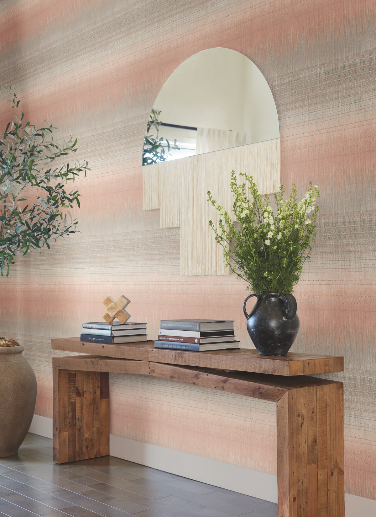 A wooden console table with books, a black vase of white flowers, and decor sits against a peach and gray Desert Textile Wallpaper by York Wallcoverings. An arched mirror and potted plant are nearby.