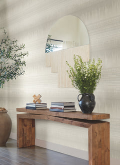 A modern, minimalist room features York Wallcoverings’ Desert Textile Wallpaper as a backdrop for a wooden console table displaying stacked books, a black vase with green foliage, and a geometric decor piece beneath a large, uniquely shaped mirror.