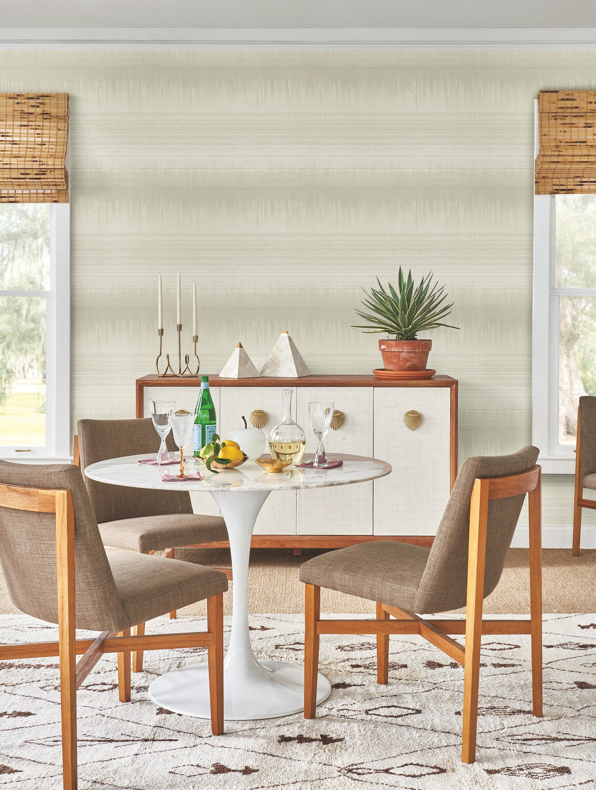 A mid-century modern dining area featuring York Wallcoverings’ Desert Textile Wallpaper, a round white table, four wooden chairs, a sideboard, potted plant, decorative objects, drinks, fruit, and large windows with woven blinds.