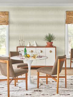 A mid-century modern dining area featuring York Wallcoverings’ Desert Textile Wallpaper, a round white table, four wooden chairs, a sideboard, potted plant, decorative objects, drinks, fruit, and large windows with woven blinds.