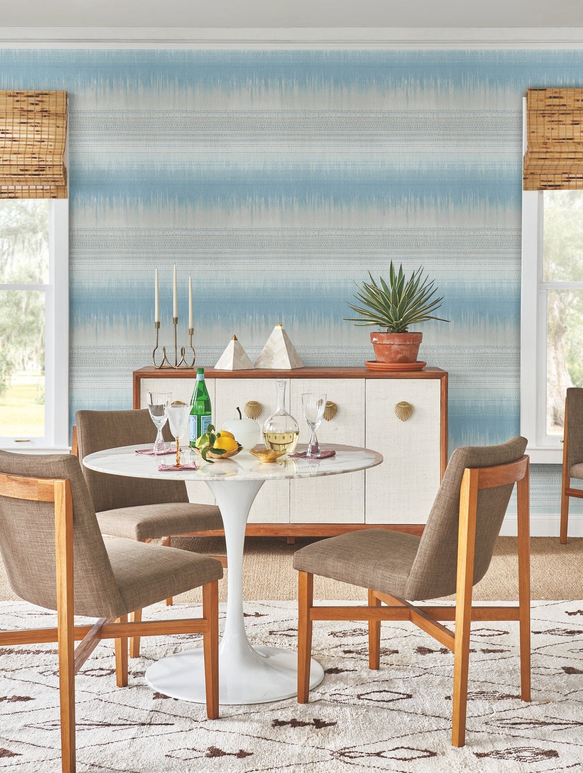 A round white table with four wooden chairs sits on a patterned rug. The table has drinks, glasses, and fruit. A credenza and plant are in the background, with the walls accented in York Wallcoverings Desert Textile Wallpaper. Two windows have bamboo shades.