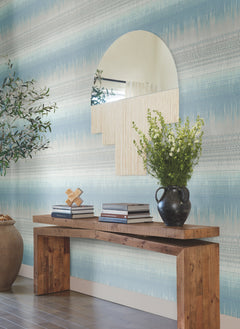 A rustic wooden console table with stacked books, a black vase of white flowers, and a decorative object sits beneath a large, uniquely shaped mirror on a wall accented with Desert Textile Wallpaper by York Wallcoverings.