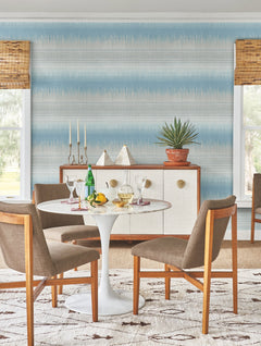 A round white table with drinks, fruit, and glasses sits with four wooden chairs. Behind, there’s a sideboard adorned with plants, decor, and a candelabra against York Wallcoverings’ Desert Textile Wallpaper—a blue patterned backdrop—framed by two windows dressed in bamboo blinds.