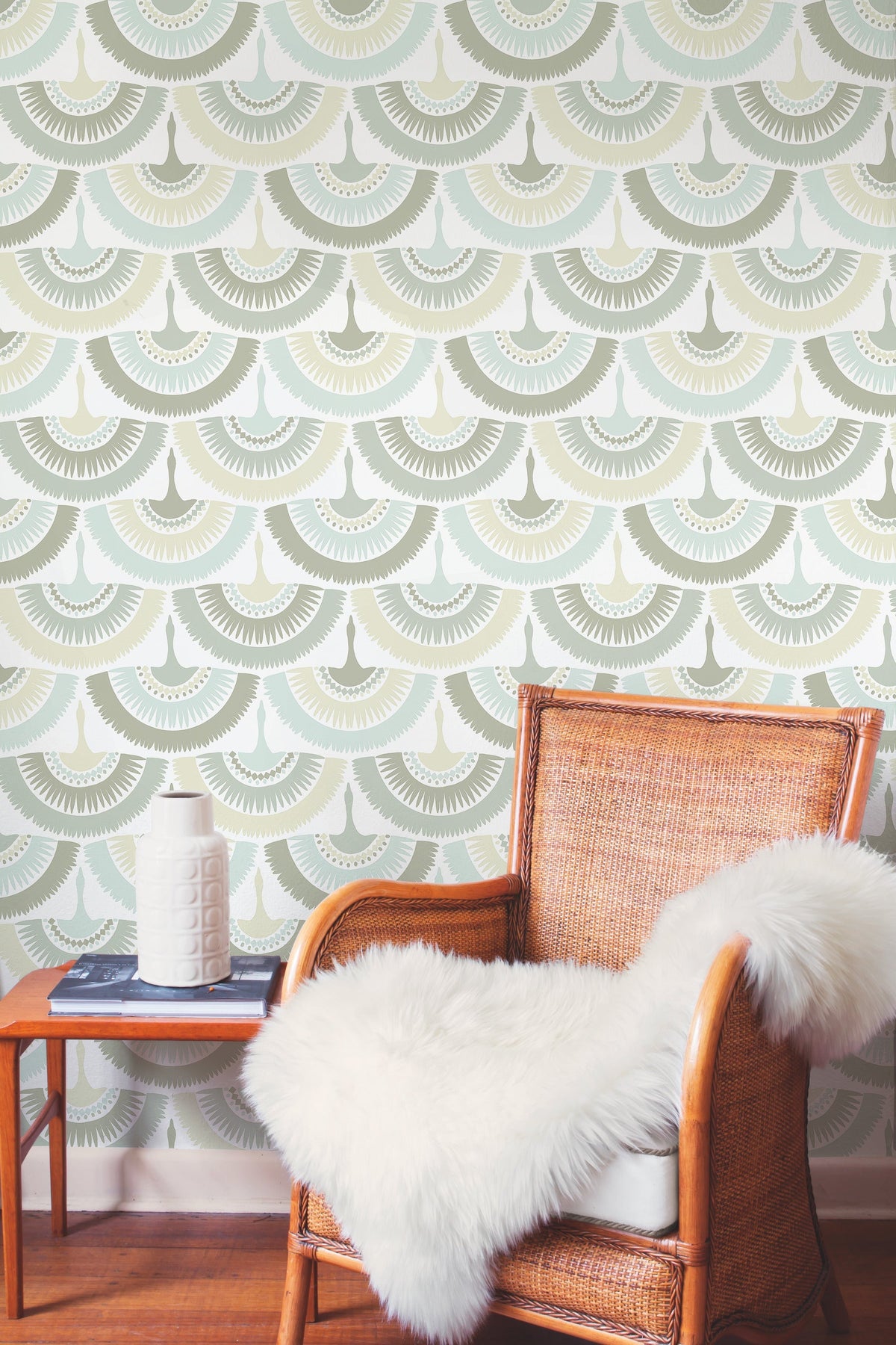 A wicker chair with a white faux fur throw sits next to a small table with a textured white vase, set against a wall adorned with York Wallcoverings’ Feather & Fringe wallpaper.