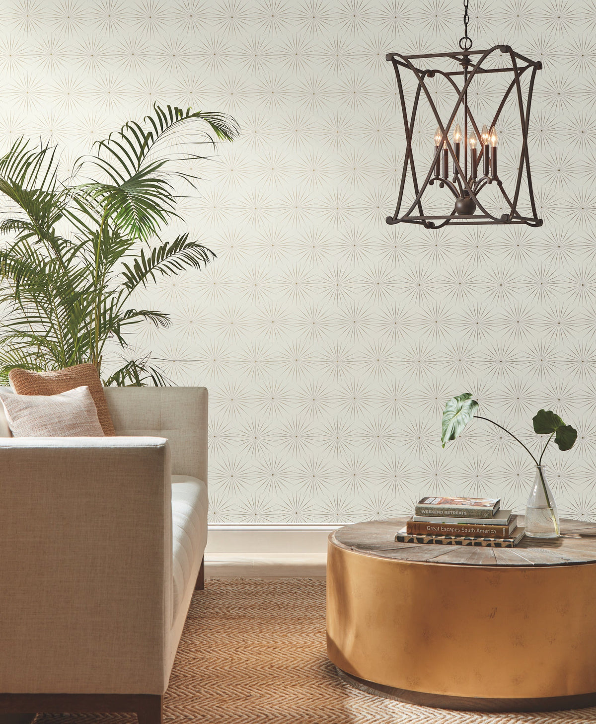 A modern living room featuring a beige sofa, a gold round coffee table, potted plants, books, and a geometric chandelier. The walls are adorned with Starlight Wallpaper by York Wallcoverings, showcasing a subtle starburst pattern. A woven rug covers the floor.