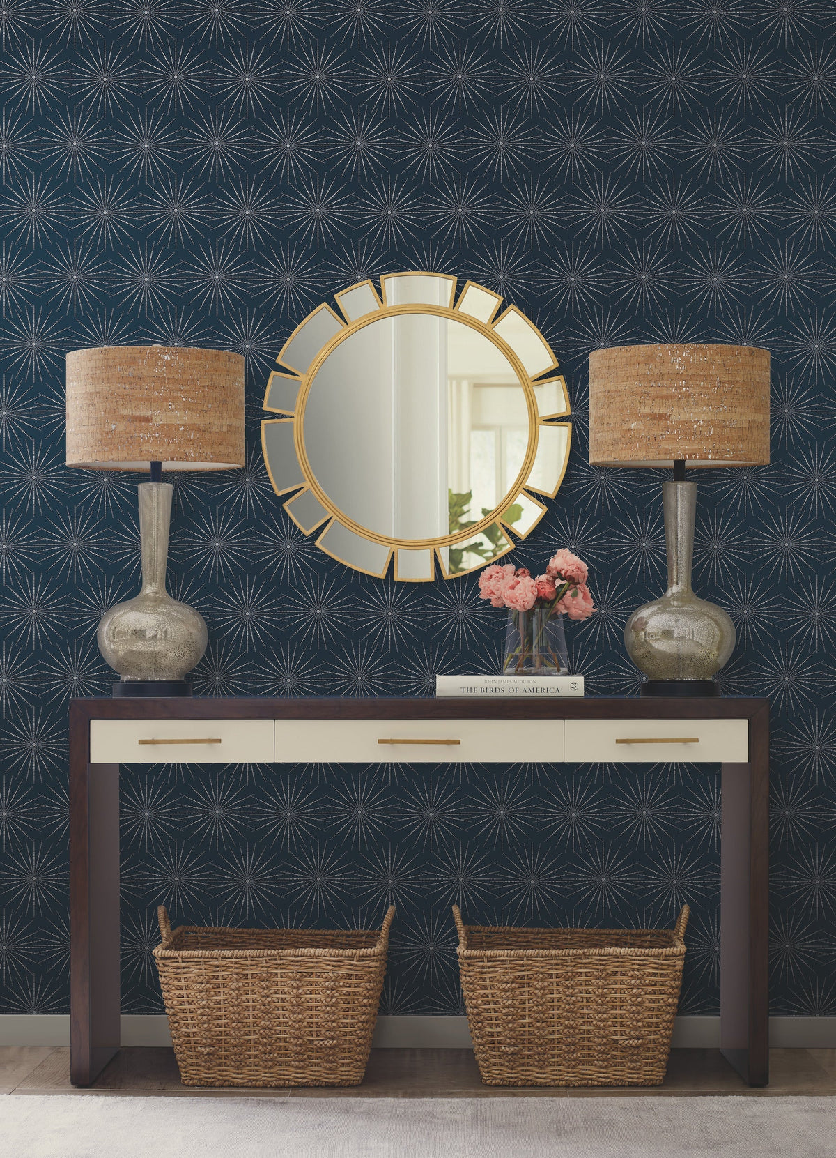 A hallway table stands against a wall adorned with York Wallcoverings’ Starlight Wallpaper, featuring two woven lamps, a gold round mirror, a vase of pink flowers, a book on the tabletop, and two wicker baskets placed underneath.