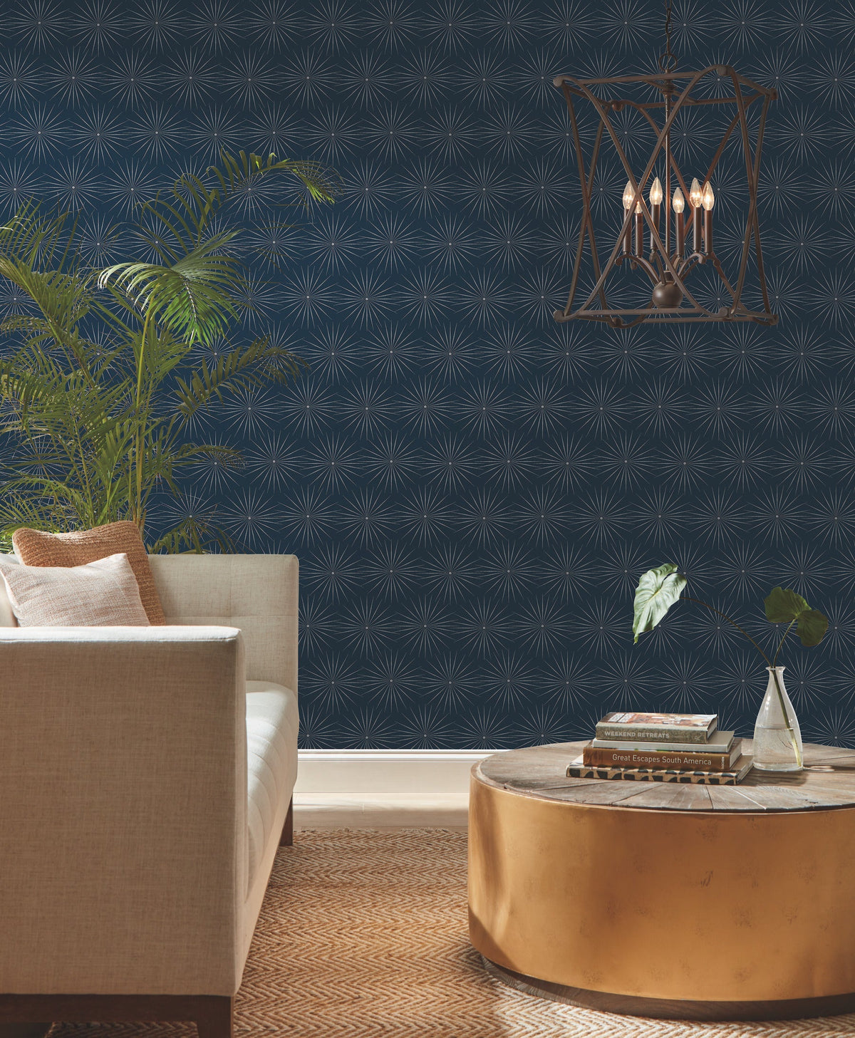 A modern living room with a beige sofa, gold round coffee table, leafy plants, Starlight Wallpaper by York Wallcoverings featuring geometric navy designs, and a hanging lantern-style chandelier over a brown patterned rug.