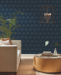 A modern living room with a beige sofa, gold round coffee table, leafy plants, Starlight Wallpaper by York Wallcoverings featuring geometric navy designs, and a hanging lantern-style chandelier over a brown patterned rug.