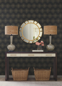 A console table with three drawers and two wicker baskets underneath is styled with two lamps, a vase of pink flowers, and topped by a circular gold mirror. The wall behind features the York Wallcoverings Starlight Wallpaper, displaying a dramatic dark geometric starburst pattern.