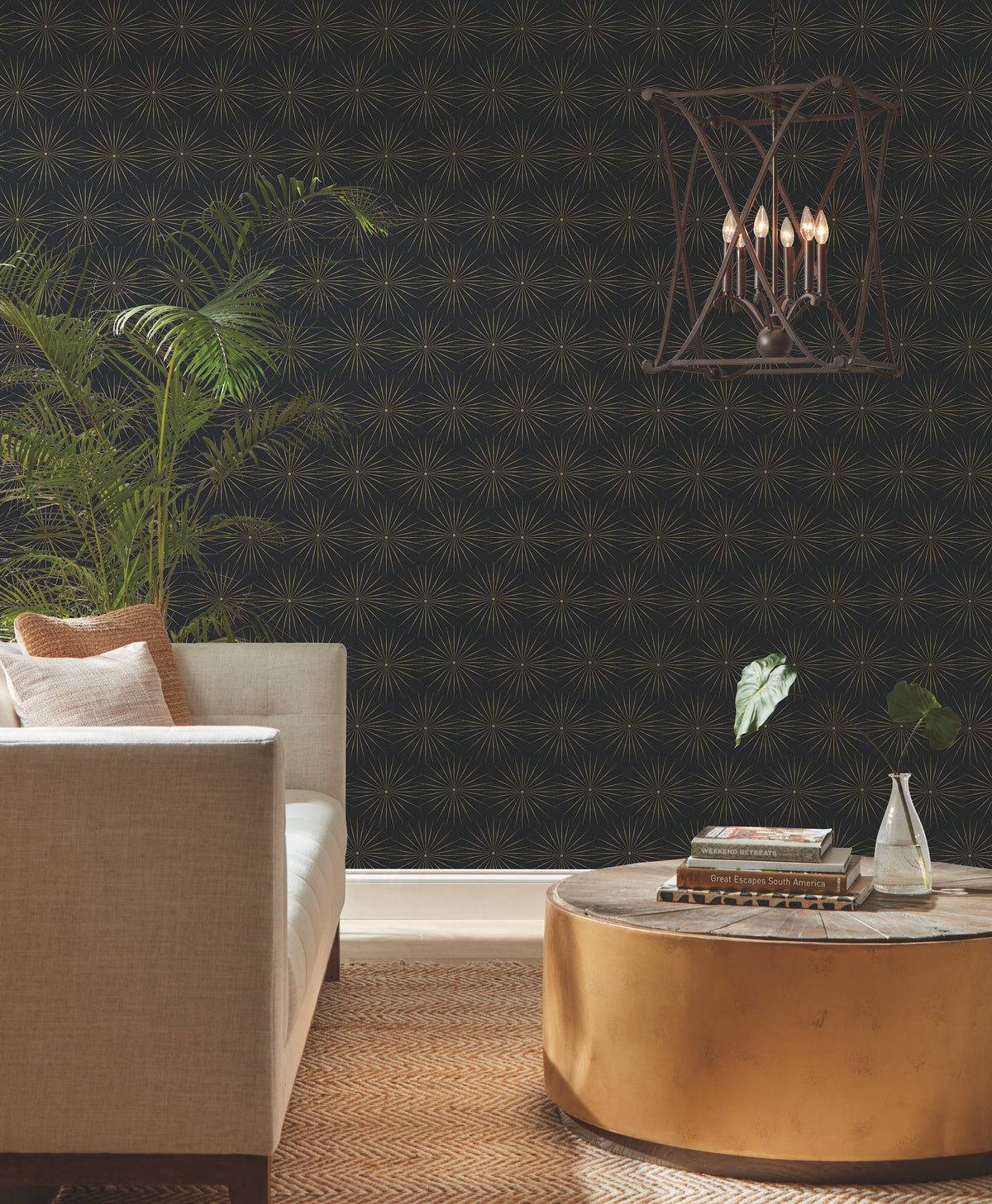 A modern living room with a beige sofa, gold round coffee table, potted plants, York Wallcoverings’ Starlight Wallpaper in black and gold, and a geometric pendant light hanging above.