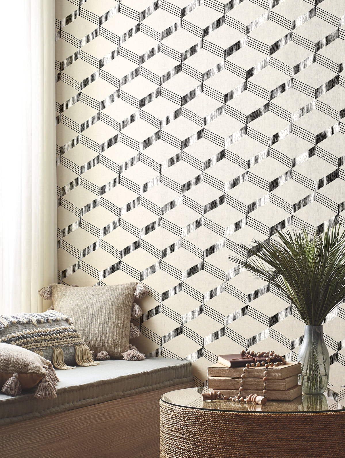 A cozy nook features beige cushions with tassels, a round wicker table topped with books and beads, and Palisades Paperweave Wallpaper by York Wallcoverings in a geometric pattern. Soft light filters through white curtains.