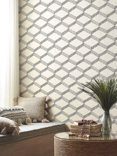 A cozy nook features beige cushions with tassels, a round wicker table topped with books and beads, and Palisades Paperweave Wallpaper by York Wallcoverings in a geometric pattern. Soft light filters through white curtains.