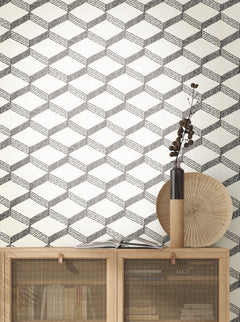 A wooden cabinet with books and decor sits against a white wall adorned with York Wallcoverings’ Palisades Paperweave Wallpaper in a black geometric cube pattern. On top are a tall vase with dried branches and a round fan-shaped decor piece.