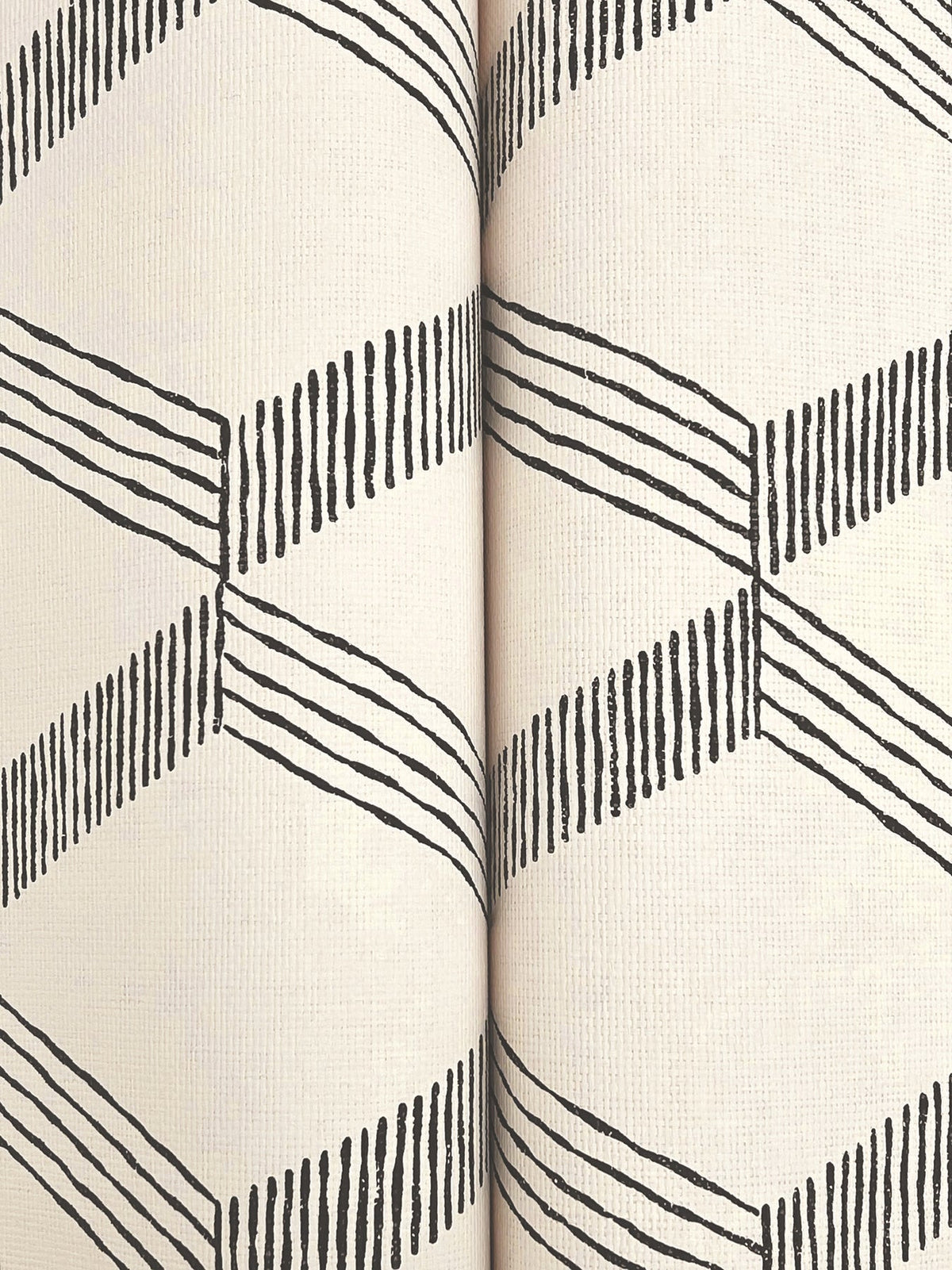 Close-up of two rolls of Palisades Paperweave Wallpaper by York Wallcoverings, featuring a cream background and a repeating black geometric zigzag pattern made from angled lines.