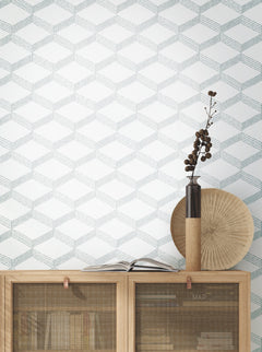 A wooden cabinet with books and decor sits against a wall covered in York Wallcoverings’ Palisades Paperweave Wallpaper, featuring a geometric pattern. On top are a tall vase with branches, a round textured plate, and an open book.