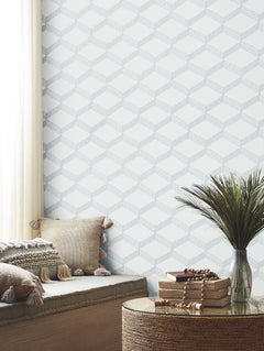 A cozy reading nook with neutral cushions, a round woven table holding books and beads, and a green plant is set against the soft gray and white geometric pattern of York Wallcoverings’ Palisades Paperweave Wallpaper. Light pours in through a sheer curtain.