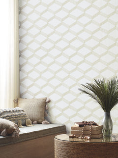 A cozy nook with beige cushions, tasseled pillows, a round wicker table topped with stacked books and a plant, set beside a window draped in sheer white curtains, features geometric-patterned Palisades Paperweave Wallpaper by York Wallcoverings.