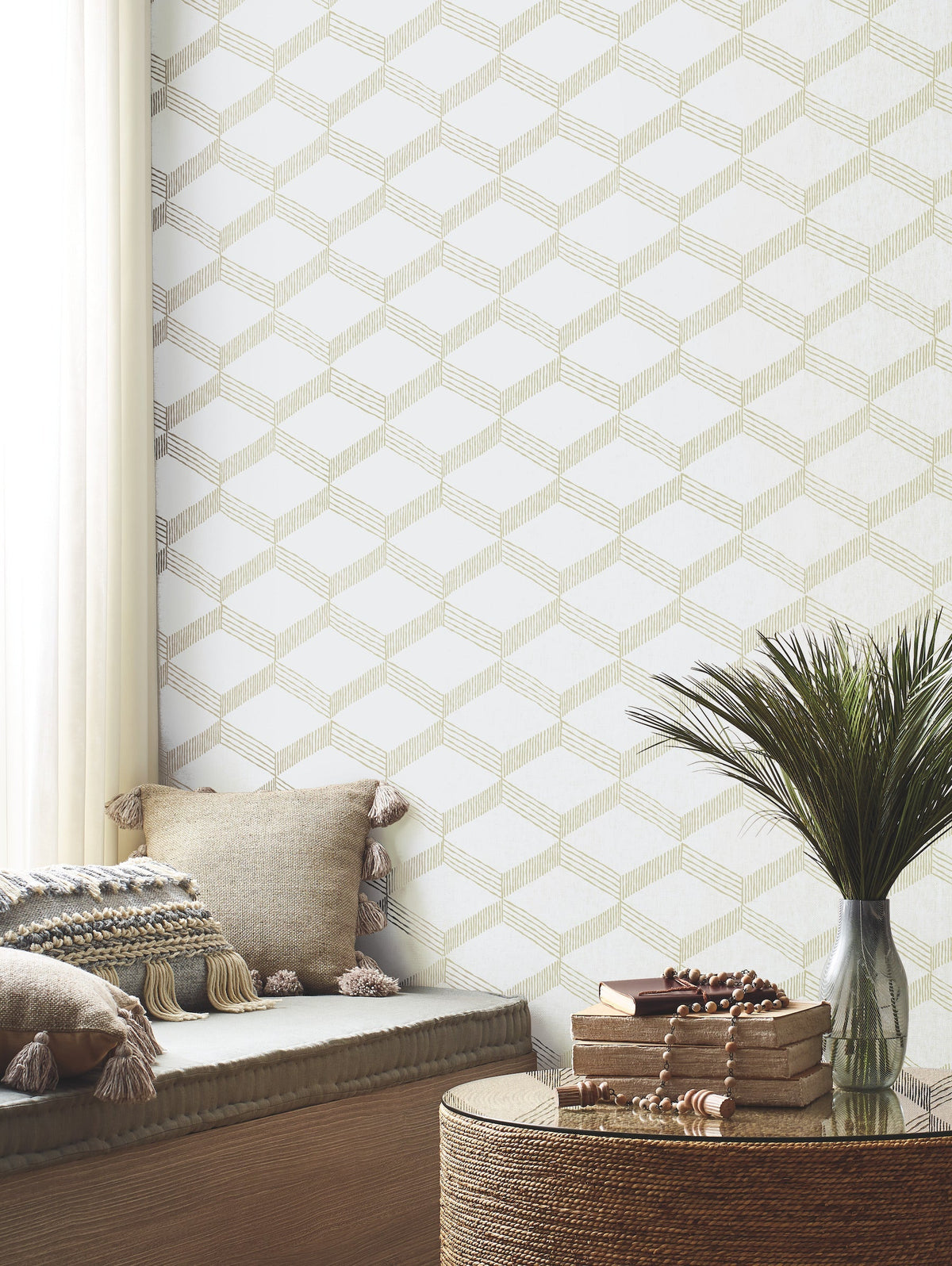A cozy nook featuring beige tasseled pillows, a small potted plant, and stacked books on a round woven table sits by a window with sheer white curtains, while the wall is adorned with York Wallcoverings’ Palisades Paperweave Wallpaper.