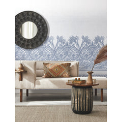 A modern living room featuring a beige sofa, a round mirror on the wall, York Wallcoverings’ Henna Wall Mural as patterned blue wallpaper, a rustic side table with books and vases, and a textured rug on the floor.