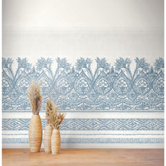 Two tall wicker vases filled with dried pampas grass stand on a wooden floor in front of a white wall adorned with the Henna Wall Mural by York Wallcoverings, featuring intricate blue floral patterns.