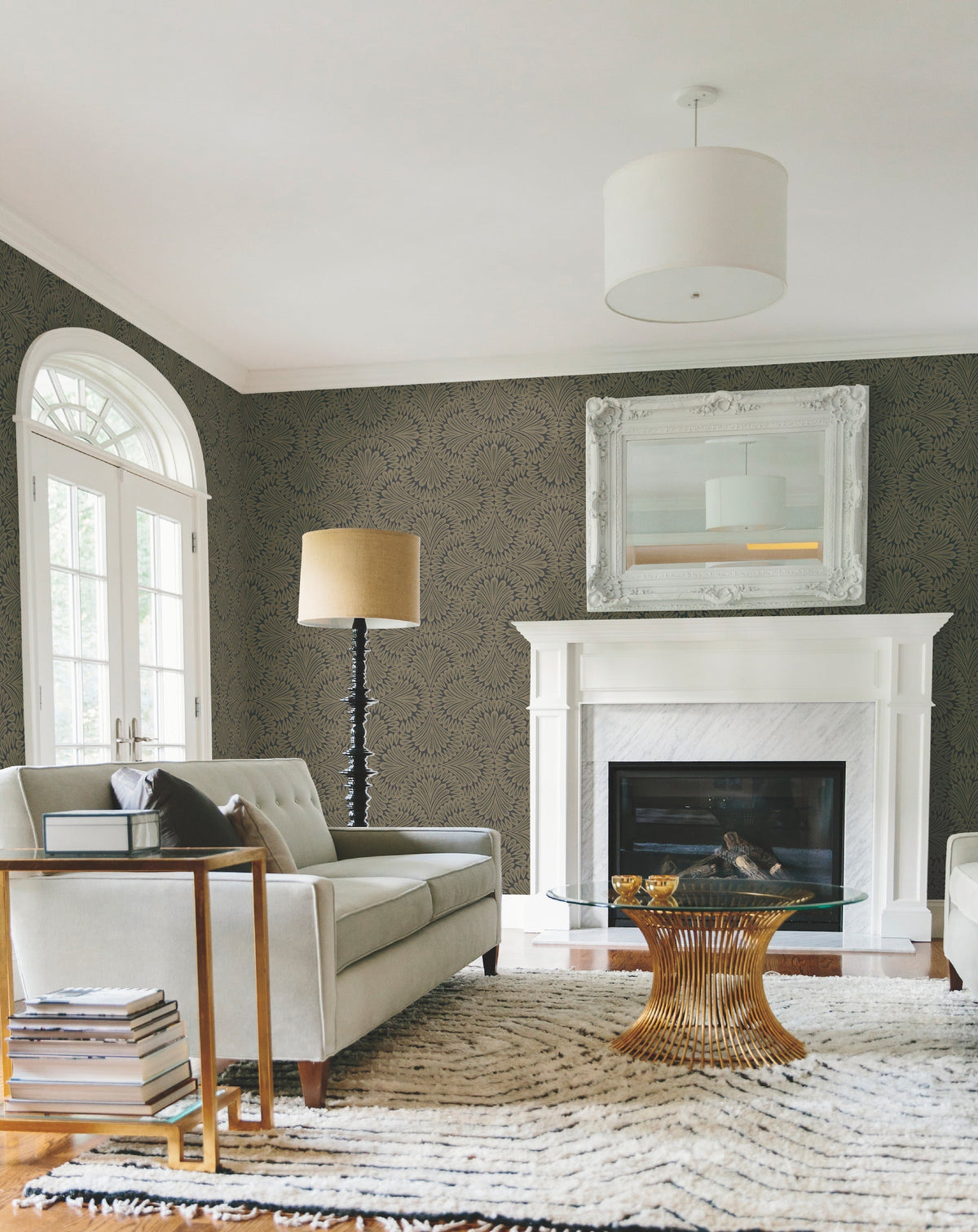 A modern living room features a gray sofa, glass coffee table, fireplace, large mirror, tall lamp, and French doors. Stacked books serve as a side table next to the sofa. The room is decorated in neutral tones with patterned Cabaret Wallpaper by York Wallcoverings.