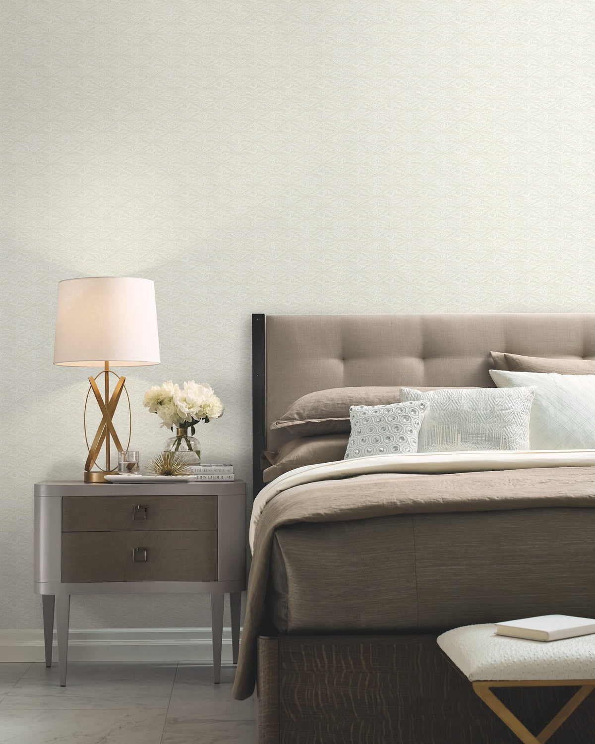 A modern bedroom featuring a tufted headboard, neutral bedding, and a gray nightstand holding a lamp, white flowers, and a book. At the foot of the bed sits a white textured bench. The walls are adorned with York Wallcoverings’ High Society Wallpaper in a light patterned design.