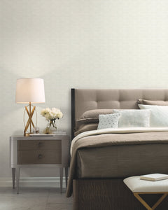 A modern bedroom featuring a tufted headboard, neutral bedding, and a gray nightstand holding a lamp, white flowers, and a book. At the foot of the bed sits a white textured bench. The walls are adorned with York Wallcoverings’ High Society Wallpaper in a light patterned design.