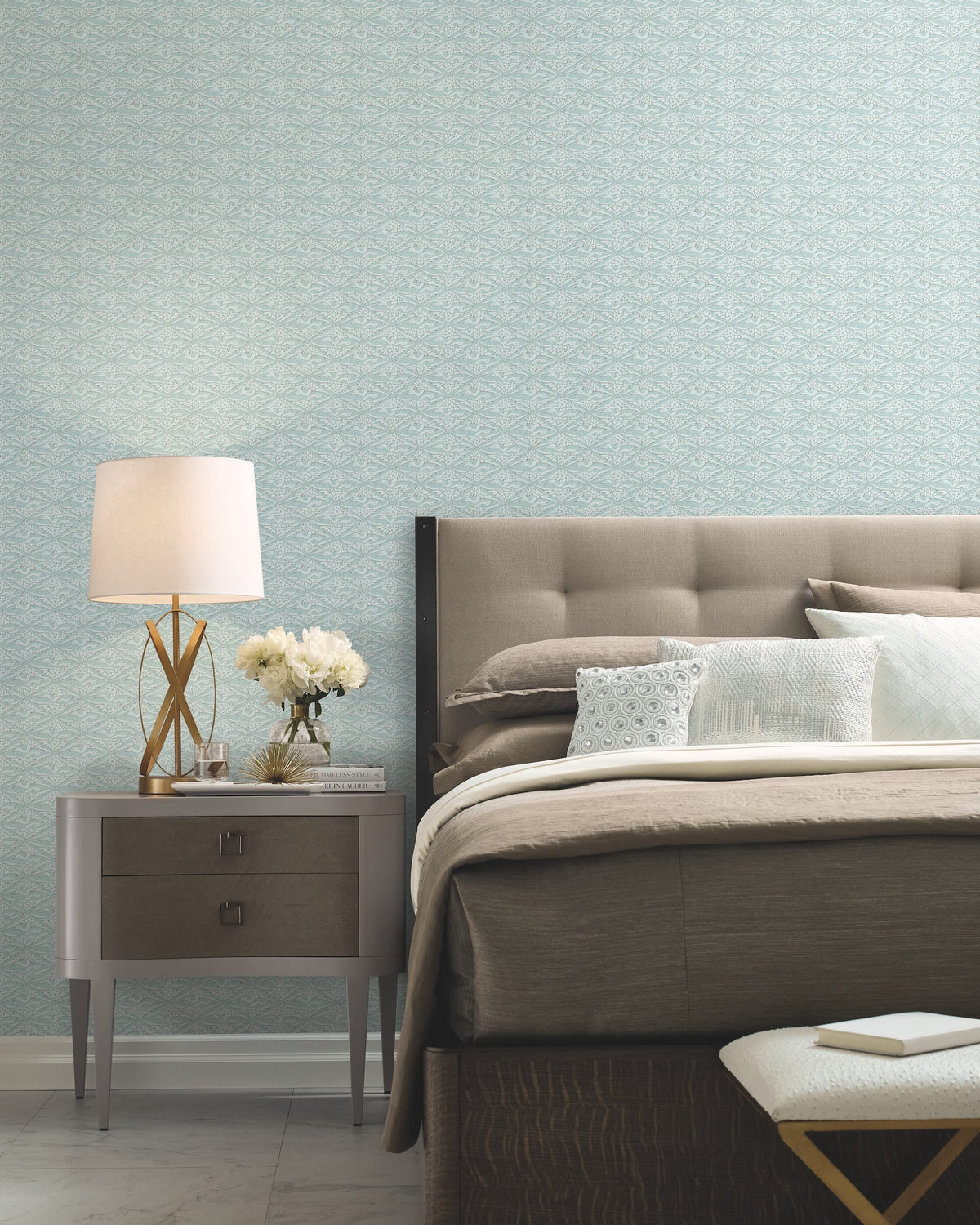 Modern bedroom with a tufted headboard, neutral bedding, and a nightstand topped with a lamp, books, and white flowers—all set against York Wallcoverings’ High Society Wallpaper in light blue.
